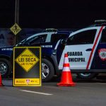 Policiais realizam uma blitz noturna com viaturas e cones em uma via urbana.