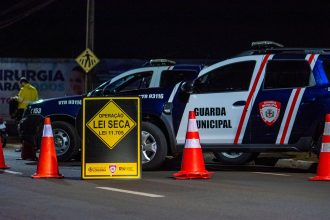 Policiais realizam uma blitz noturna com viaturas e cones em uma via urbana.