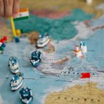 Mapa-múndi com pequenos navios e bandeiras indicando diferentes localidades e rotas comerciais globais.