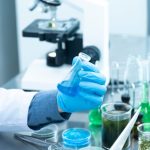 Cientista usando luvas analisa um líquido azul em laboratório, diante de um microscópio e materiais de pesquisa.