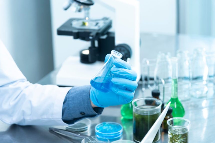 Cientista usando luvas analisa um líquido azul em laboratório, diante de um microscópio e materiais de pesquisa.