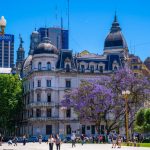 Vista urbana da Plaza de Mayo, em Buenos Aires, com edifícios históricos e árvores floridas.