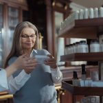 Idosa recebe orientação de uma farmacêutica em uma farmácia, diante de prateleiras com produtos.