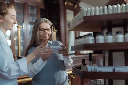 Idosa recebe orientação de uma farmacêutica em uma farmácia, diante de prateleiras com produtos.